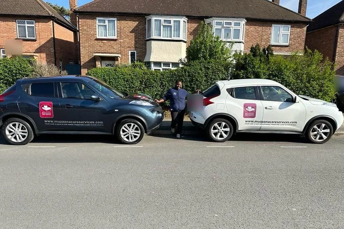 A Muzaana carer standing between two branded company cars parked on a residential street.