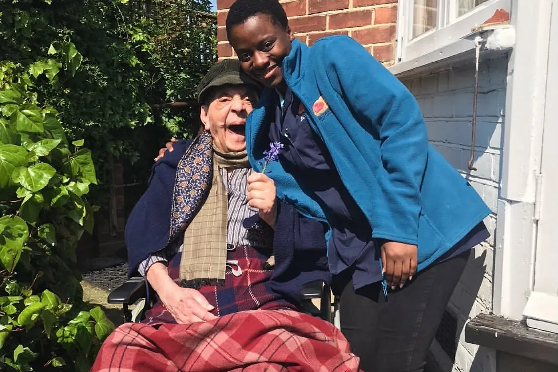 A Muzaana carer leaning in beside someone seated in a wheelchair outside a house, holding a sprig of lavender.