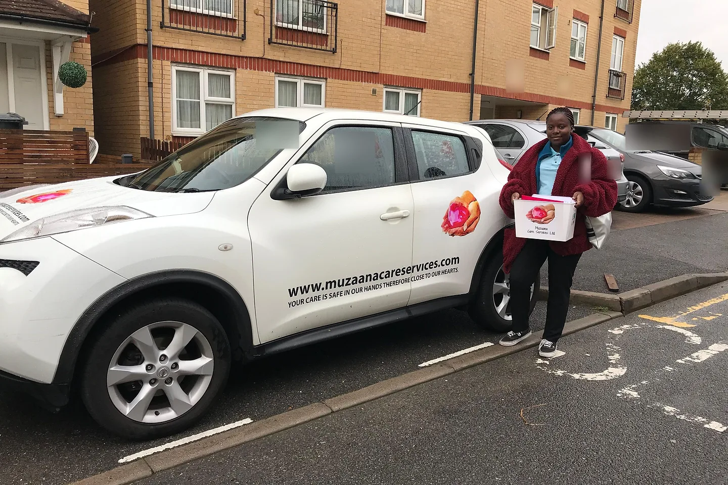 A Muzaana carer carrying a box of care records beside the branded company car outside a block of flats.