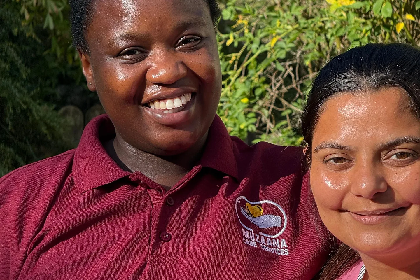 Two Muzaana staff standing together outdoors in front of a hedge, smiling in the sunshine.