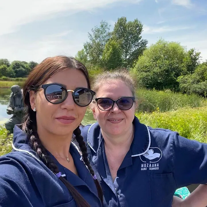 Two Muzaana carers in navy uniform outdoors beside a lake on a sunny day.