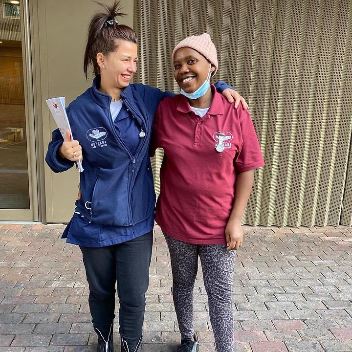 Two Muzaana carers in branded uniform outside a building, arms around each other, laughing.