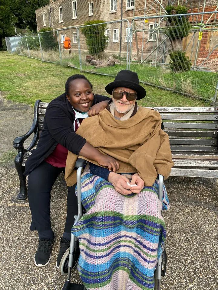 A Muzaana carer sitting on a park bench beside a man in a wheelchair, both smiling.