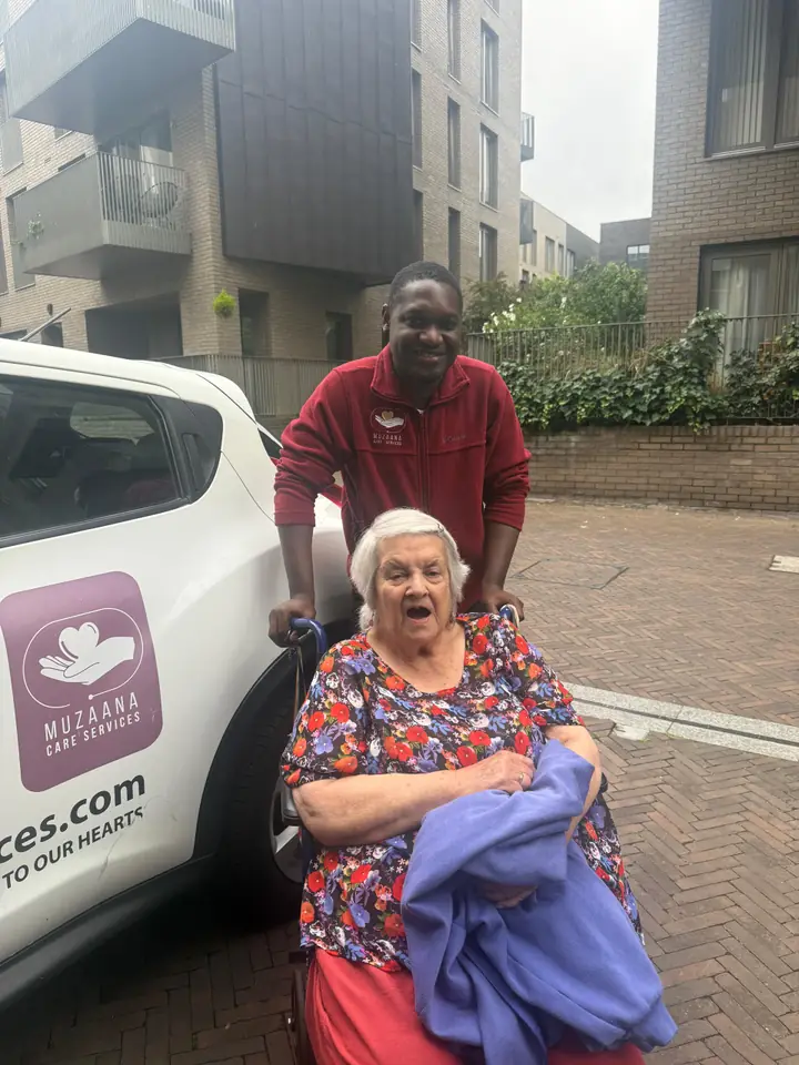 A Muzaana carer standing behind a woman in a wheelchair beside a Muzaana Care Services car.