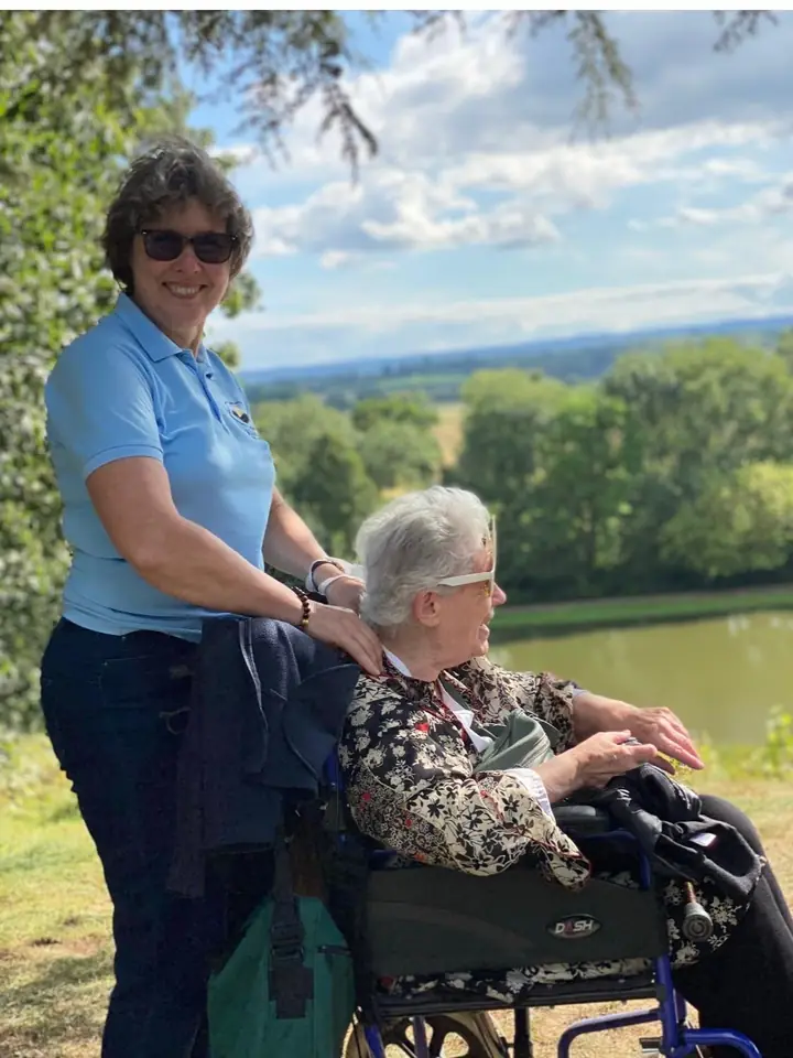 A Muzaana carer standing with a woman in a wheelchair beside a lake on a sunny day.