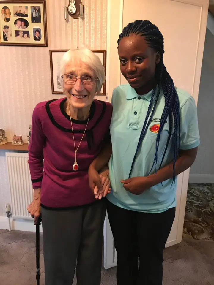 A Muzaana carer in uniform standing arm in arm with a woman in her living room.