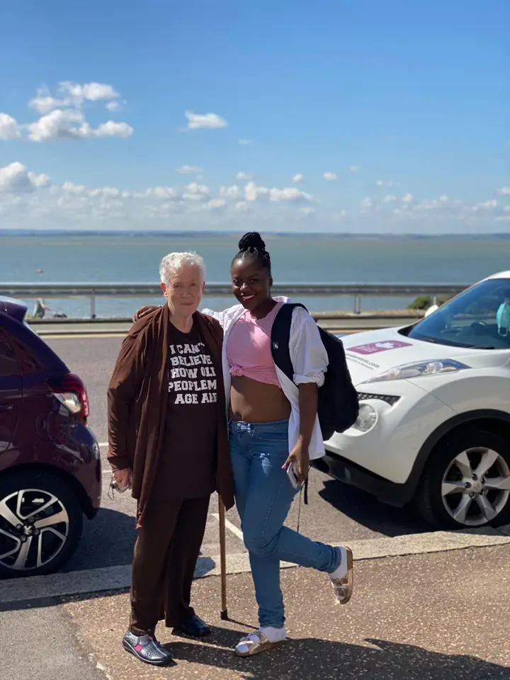 A Muzaana carer standing with a woman on a seafront, both smiling at the camera.