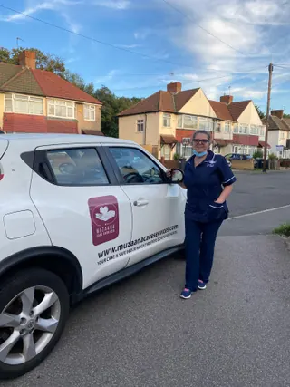 A Muzaana carer smiling beside the branded company car on a suburban street.