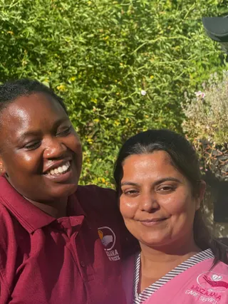 Two women in Muzaana uniforms, one maroon and one pink, smiling together in the sunshine.